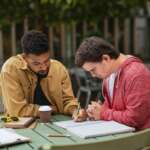 Young man with Down syndrome with his mentoring friend sitting outdoors in cafe and studying