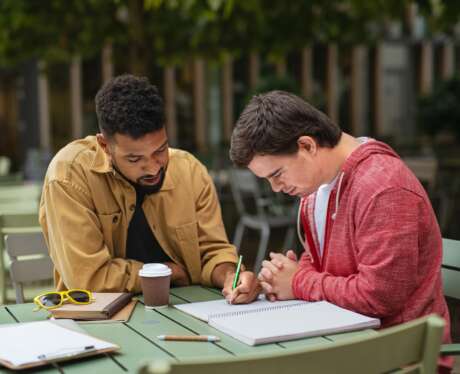 Young man with Down syndrome with his mentoring friend sitting outdoors in cafe and studying