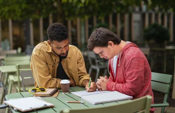 Young man with Down syndrome with his mentoring friend sitting outdoors in cafe and studying